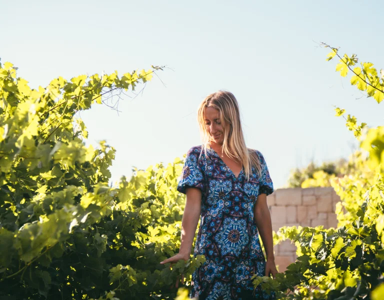 A woman immersed in sunlit vines, gently touching the leaves, wearing a patterned summer dress.