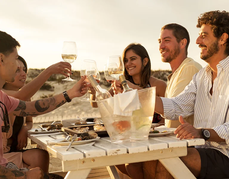 Group of friends enjoying seafood dinner with wine on a sandy beach in Portugal at sunset.