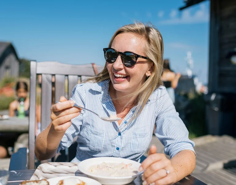 A woman in a striped shirt enjoys a bowl of soup outdoors, with blue skies and a casual dining atmosphere around her.