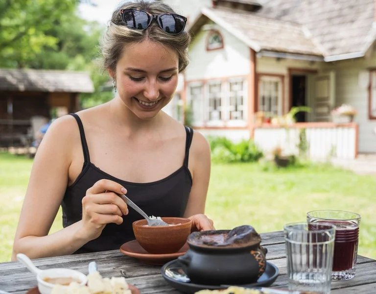 Woman enjoying traditional Seto food outdoors at Taarka Taro KöögiKono restaurant in Estonia, with rustic farm buildings in the background.