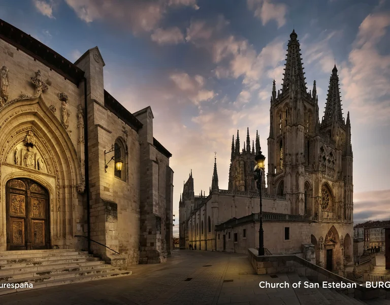 Narrow night view of Burgos Cathedral illuminated against a clear sky with scattered clouds.