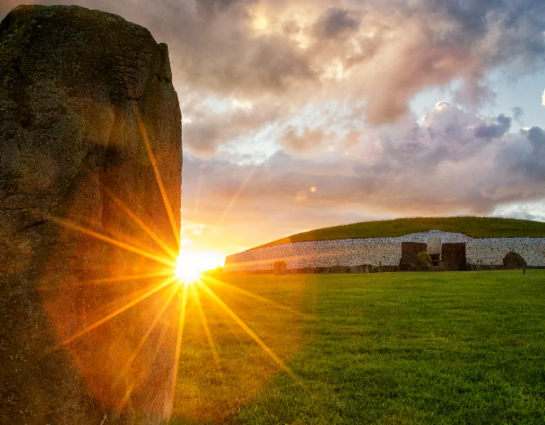 Aerial view of Newgrange, the ancient Neolithic monument in County Meath, Ireland, surrounded by green fields, bathed in warm sunset light.