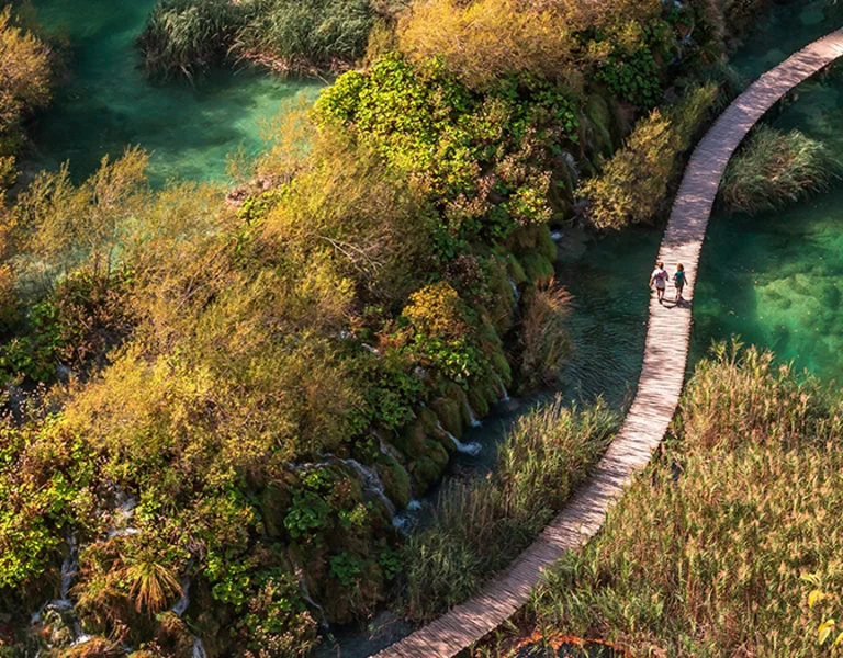Aerial view of a wooden boardwalk winding through emerald lakes and lush vegetation at Plitvice Lakes National Park, Croatia, with two visitors walking along the path.