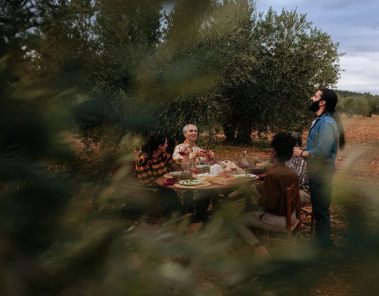 A group of friends sharing a meal outdoors among olive trees, enjoying local food and wine at a rustic wooden table in the countryside, evoking Mediterranean hospitality and slow travel.