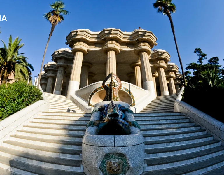 Wide-angle view of the grand staircase and mosaic dragon fountain at Park Güell in Barcelona, Spain, surrounded by palm trees and greenery.