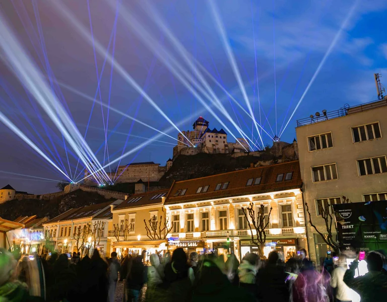 Spectacular night view of Trenčín Castle illuminated by white and blue laser beams during a public show, with a crowd watching below.