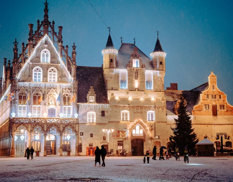 Historic center of Mechelen, Belgium, covered in snow — medieval buildings, cobblestone streets, and a church tower under a winter sky.