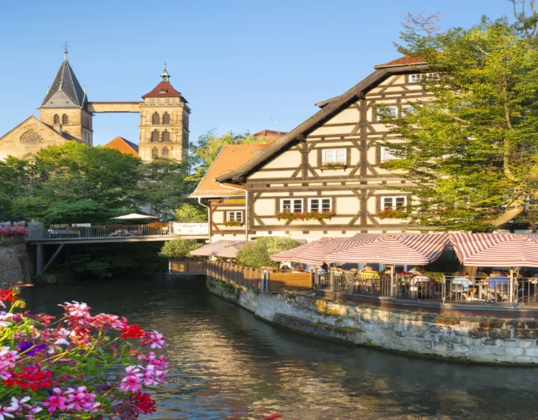Half-timbered houses by a river with pink flowers and a church in the background under blue sky.