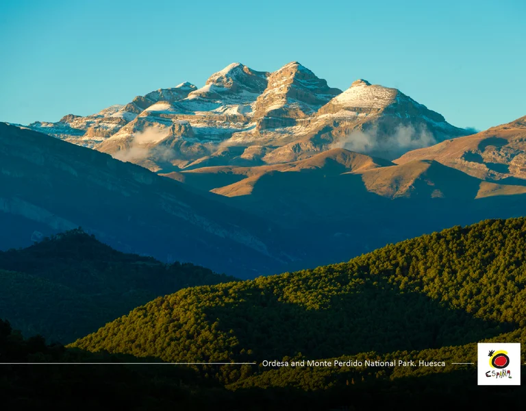 Snow-capped mountains with green forested hills in sunlight.
