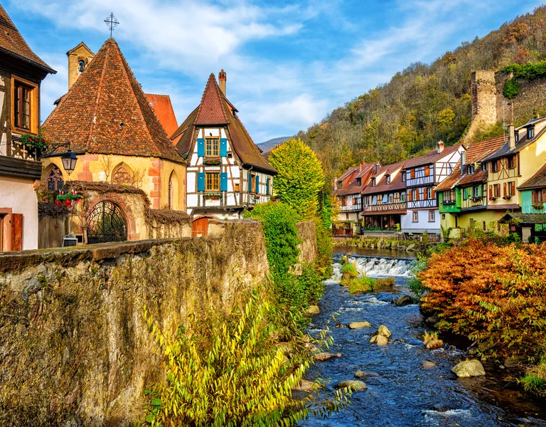 Colorful houses line a river in a quaint village with a hillside and castle ruins in the background.