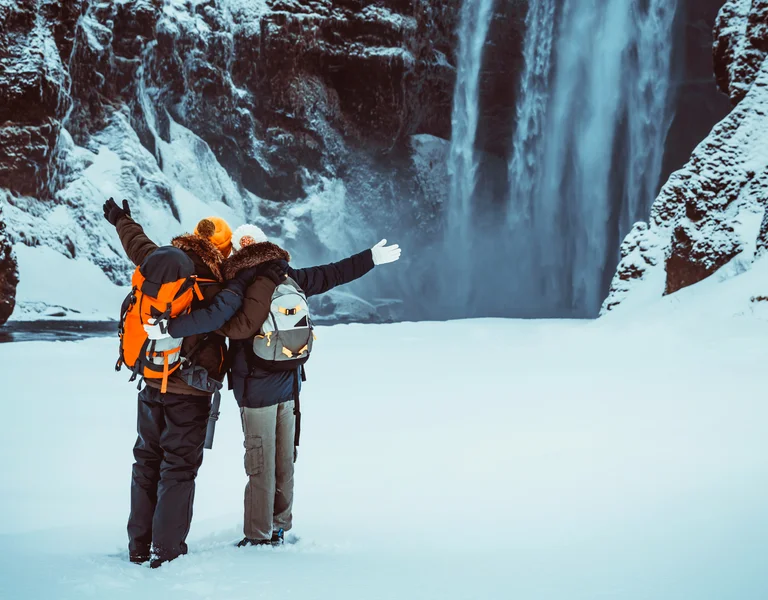 Two people in winter coats stand on snow, facing a large icy waterfall with arms raised.