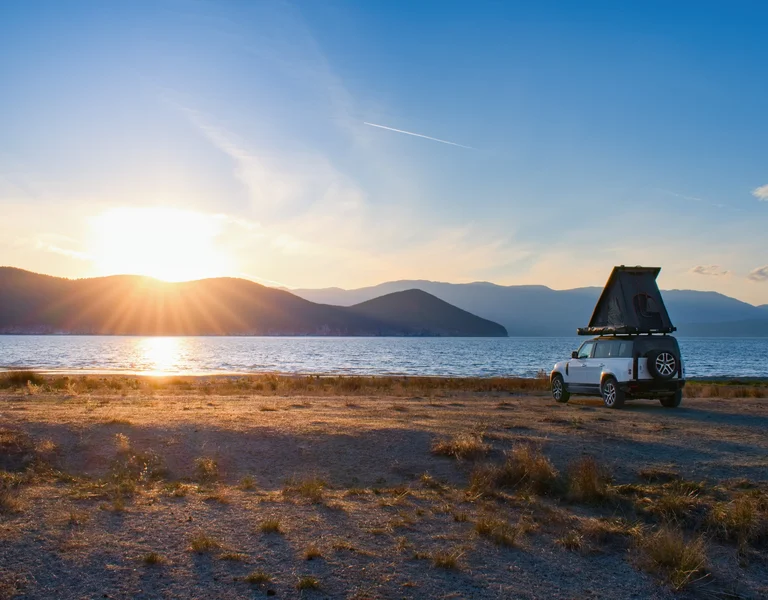 Sunset over a lake with a car and rooftop tent parked on the shore.