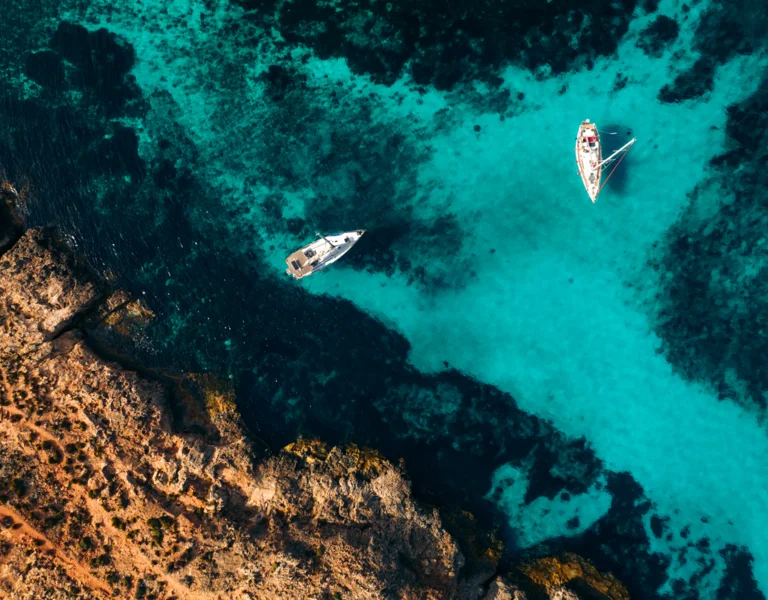 Two sailboats in clear turquoise water near a rocky coastline, photographed from above.