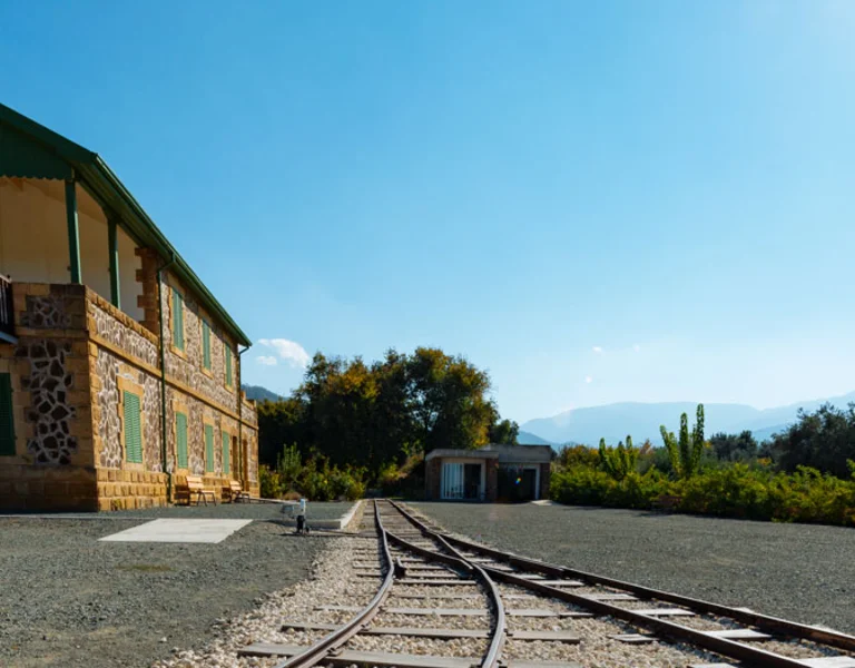 Old train station with tracks, stone building, and clear blue sky.