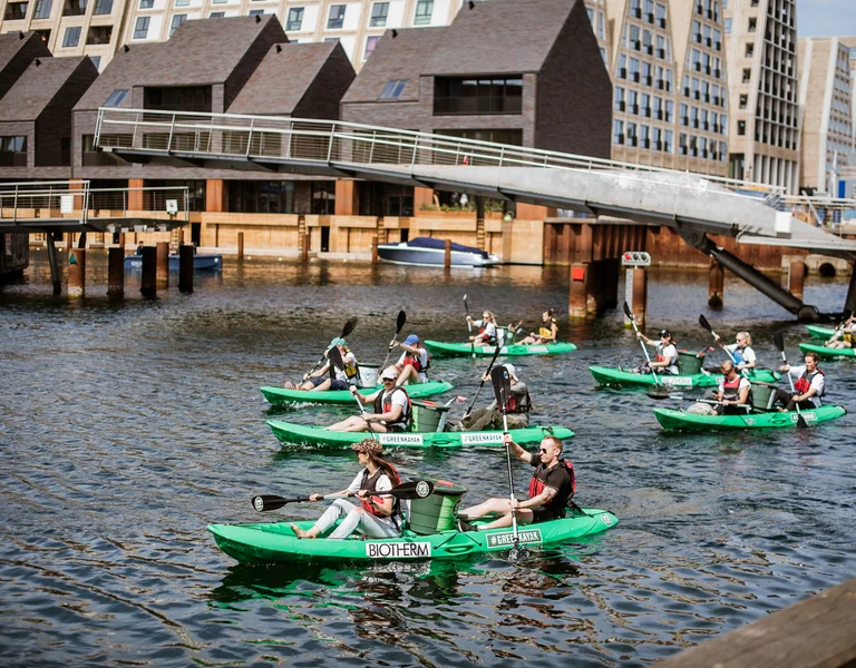 People kayaking in green boats on a city canal with modern buildings in the background.