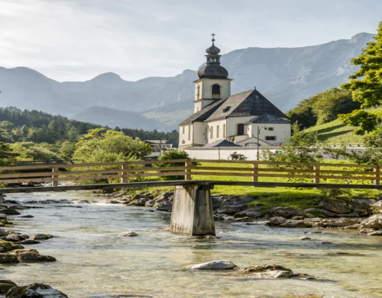 Church by a wooden bridge over a clear stream, set against mountains and lush greenery.