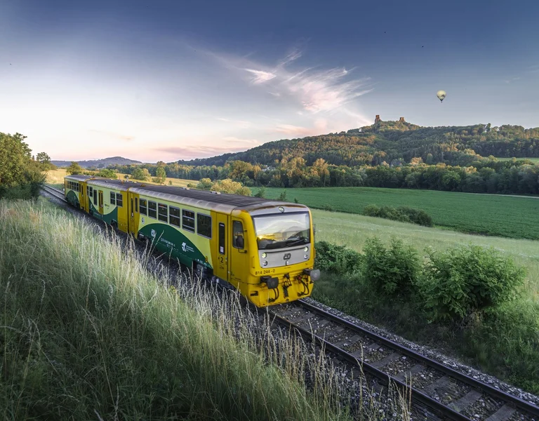 Yellow and green train traveling through grassy countryside at sunset, with hills in the background.
