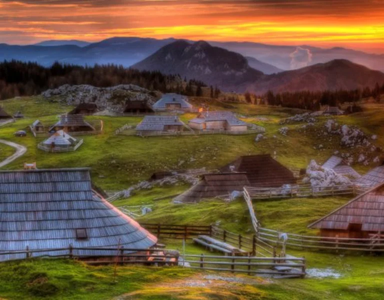 Wooden huts on grassy hills at sunset with mountains in the background.