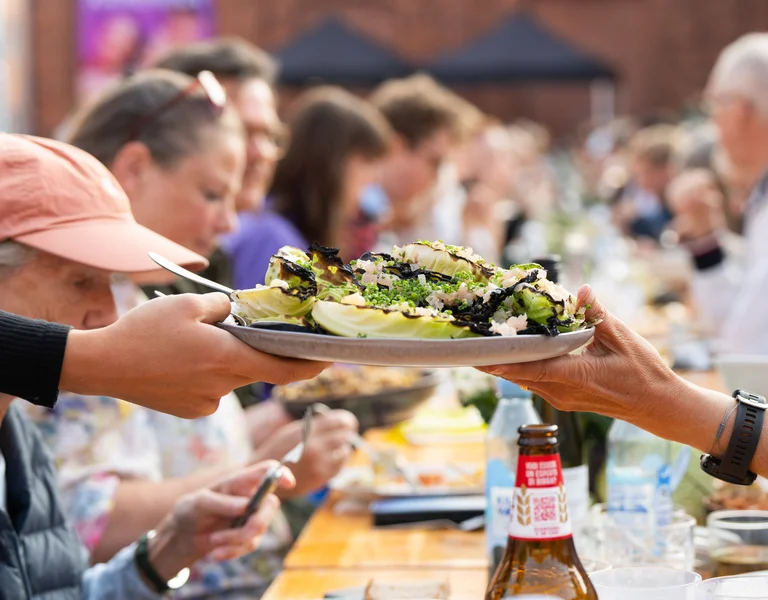 People sharing salad at a long outdoor dining table, with food and drinks.