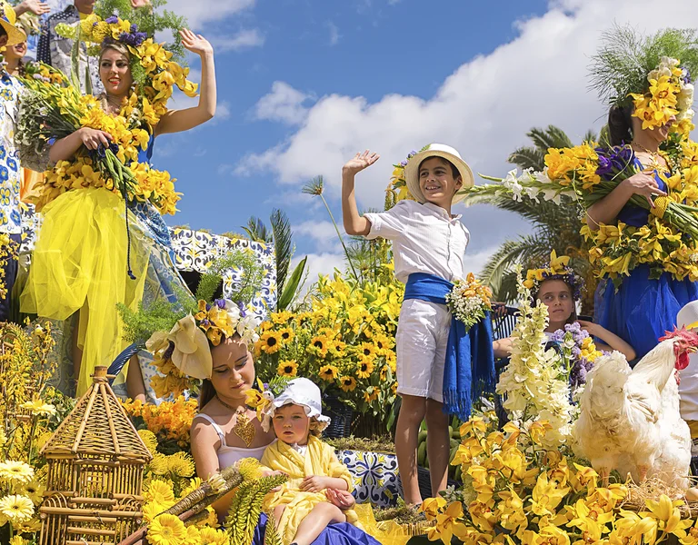 People in bright costumes on a colorful flower-covered parade float under a blue sky.