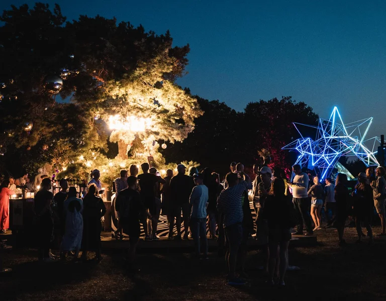 Crowd gathered outdoors at night with illuminated trees and glowing decorations.