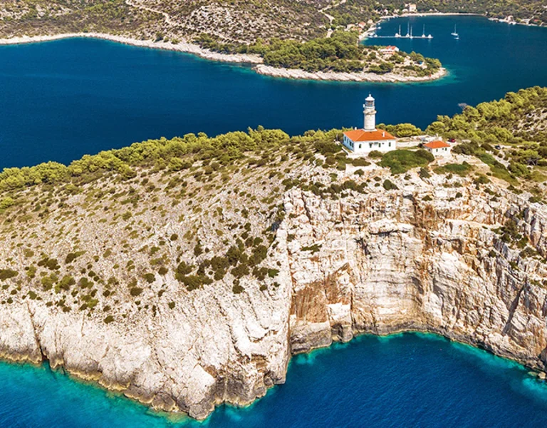 Rocky coastline with a white lighthouse surrounded by blue sea and green trees.