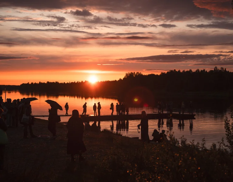 People standing by a river at sunset, silhouettes reflected on the water.