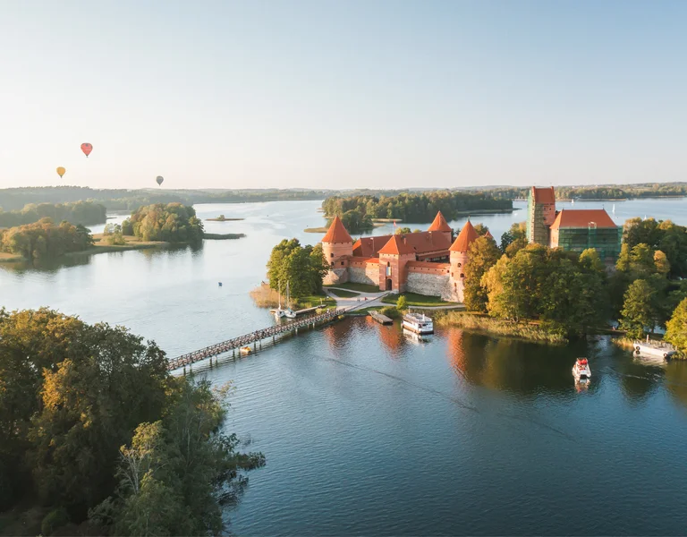 Island castle with red roofs surrounded by water, trees, and hot air balloons in the sky.