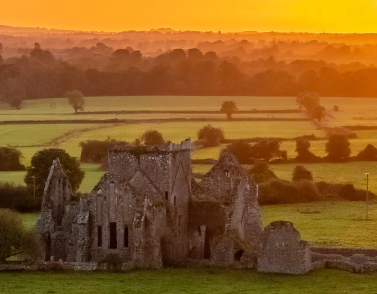 Ancient stone ruins in a green field at sunset with an orange sky.