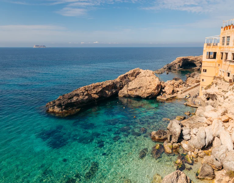 Clear blue sea with rocky coastline and a building overlooking the water.