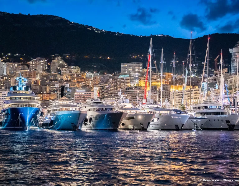 Luxury yachts docked at a marina, city lights glowing at dusk with hills in the background.
