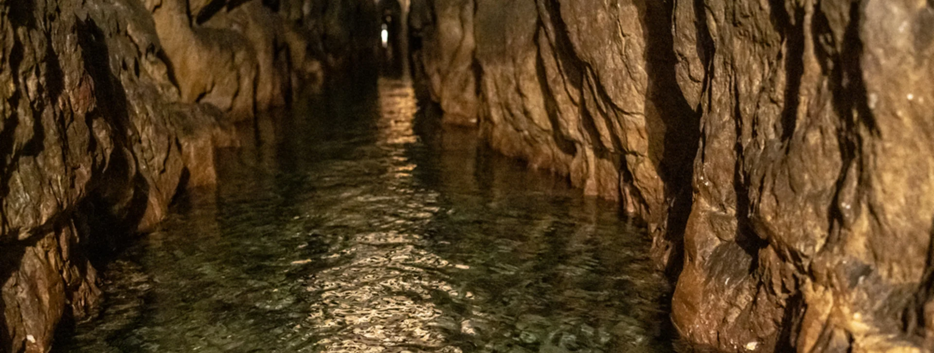 Dimly lit rock tunnel with water on the floor and distant light at the end.