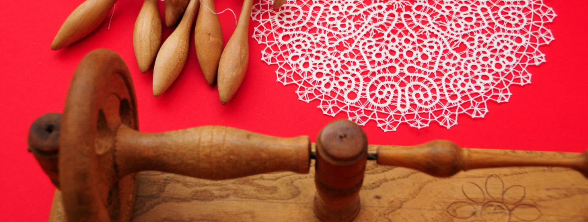 A wooden lace-making tool rests beside delicate lace, with several bobbins arranged against a vibrant red background.