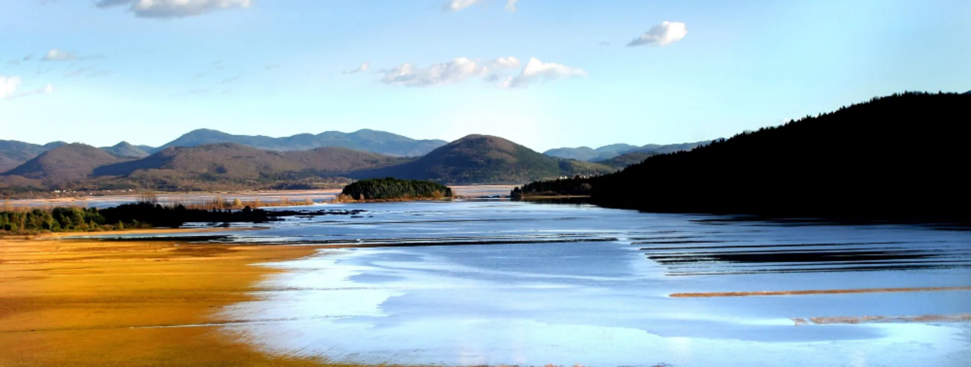 Vast landscape featuring a calm lake surrounded by rolling hills and vibrant autumn foliage under a clear blue sky with scattered clouds.