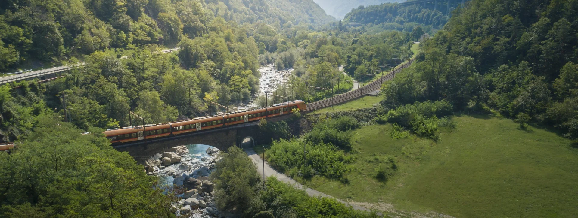 A scenic view of a train crossing a stone bridge over a river, surrounded by lush green mountains and winding railway tracks.