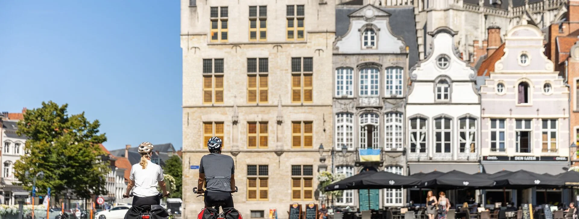 Two cyclists view historic buildings in a vibrant square on a sunny day, with cafés and greenery in the background.