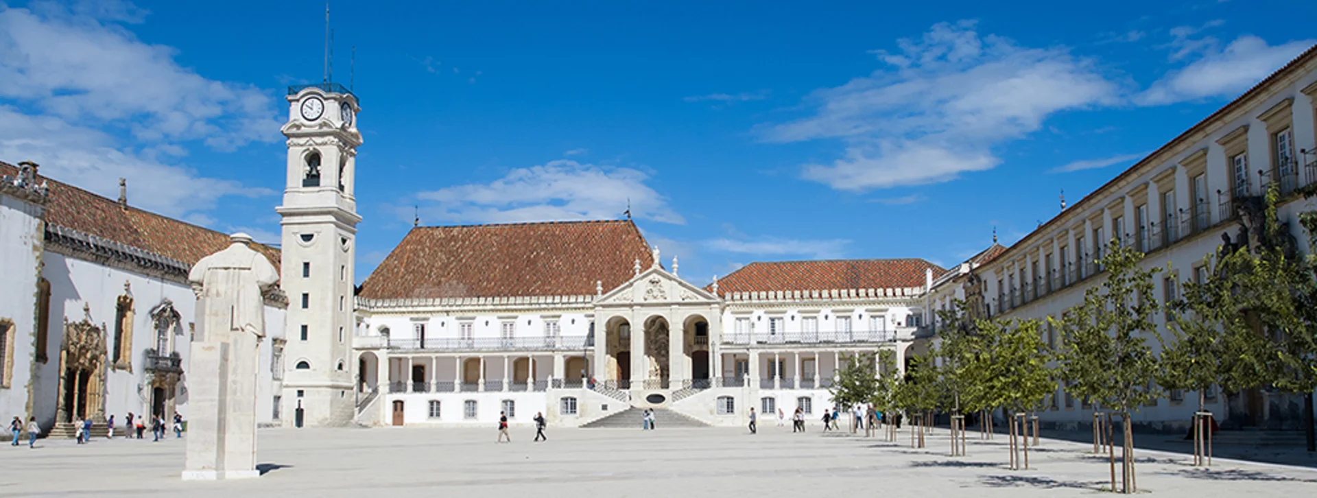 A sunlit university courtyard featuring historical buildings, a clock tower, and visitors strolling among trees under a blue sky.