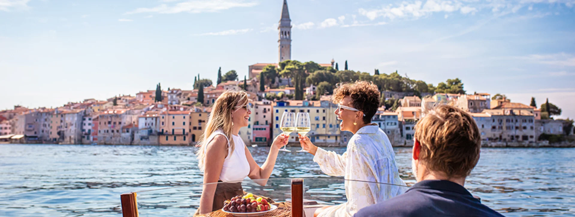 A couple cheers with glasses on a boat, with a picturesque coastal town and a bell tower in the background. A basket of grapes sits nearby.