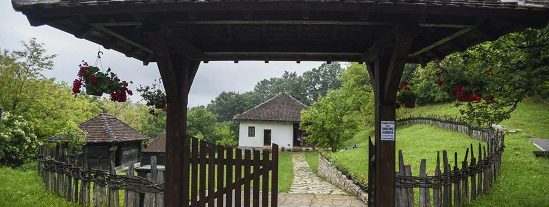 Traditional rural house in Mionica, Serbia, with a wooden gate, stone walkway, and lush green surroundings on a rainy day.