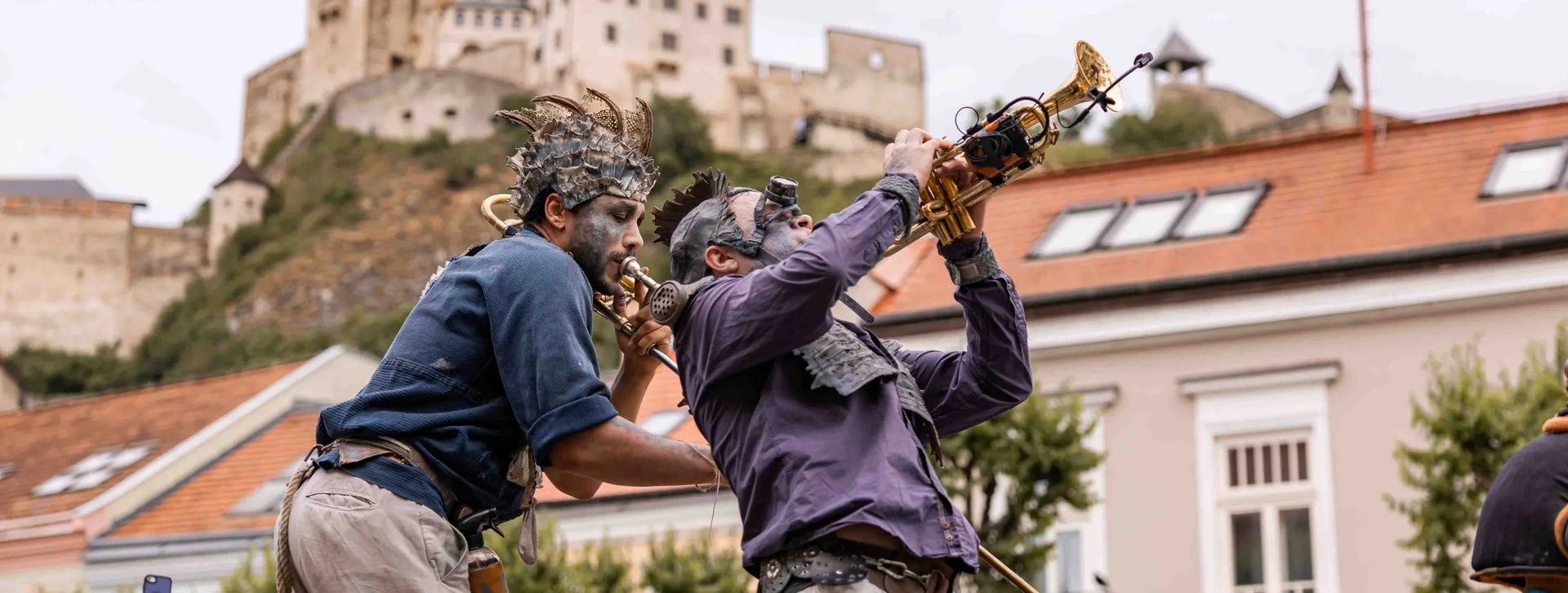 Two street performers in steampunk-style costumes playing modified brass instruments, with Trenčín Castle on the hill behind them.