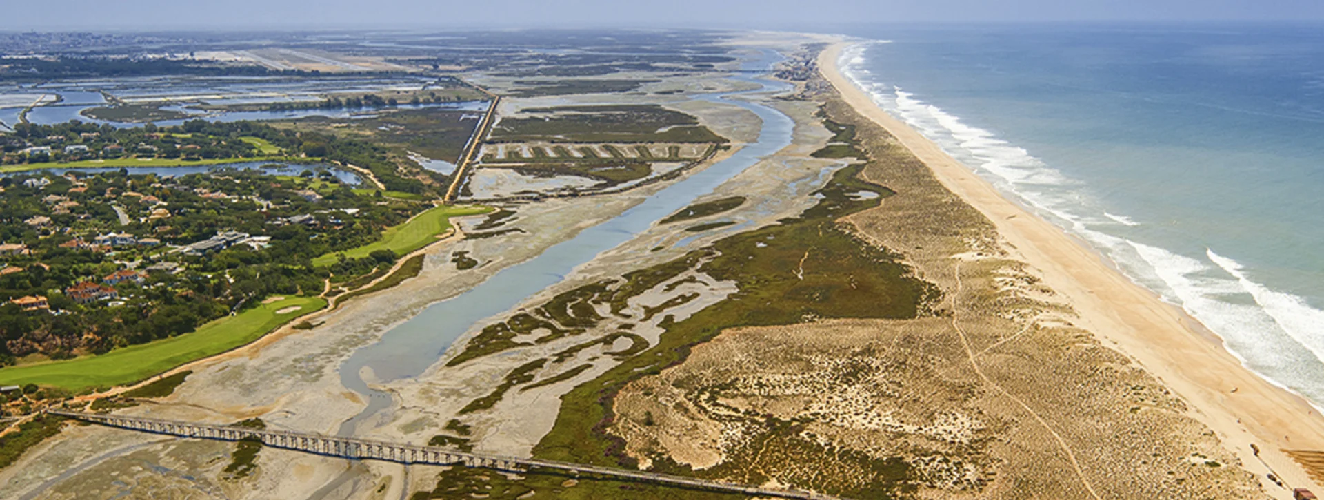 Aerial view of Quinta do Lago resort and the Ria Formosa coastal lagoon in the Algarve, Portugal, showing sandy dunes, salt‑marsh channels, and a wooden boardwalk bridge.