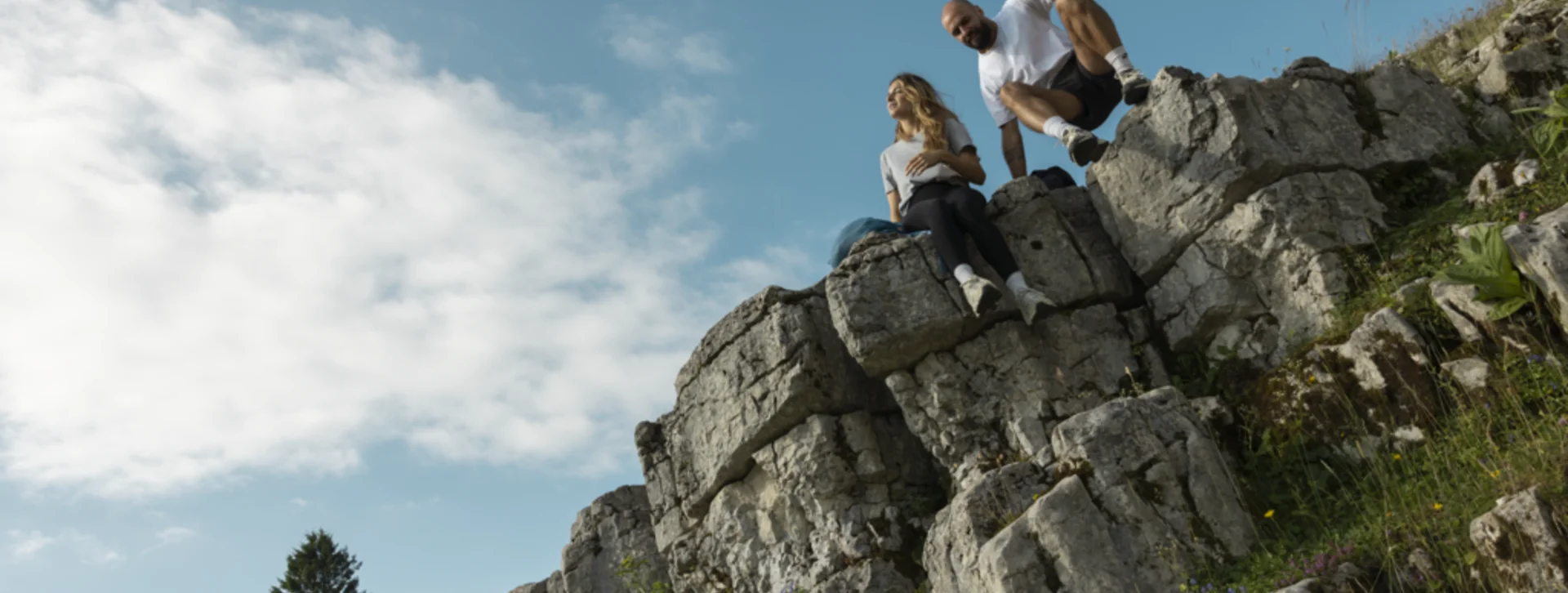 Two people sitting on rocky cliff under a bright blue sky with scattered clouds.