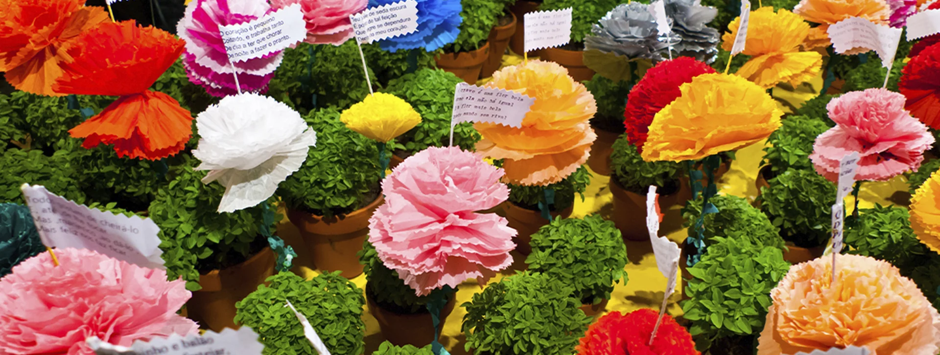 Colorful artificial flowers and green shrubs in small pots, each with a white label.
