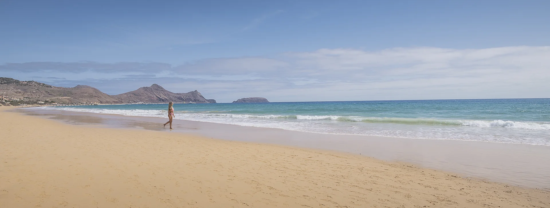 Person walking on a wide sandy beach with blue sky and gentle waves.