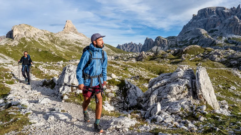 A man hiking in the dolomites in Italy. It has mountains behind and another hiker with him.