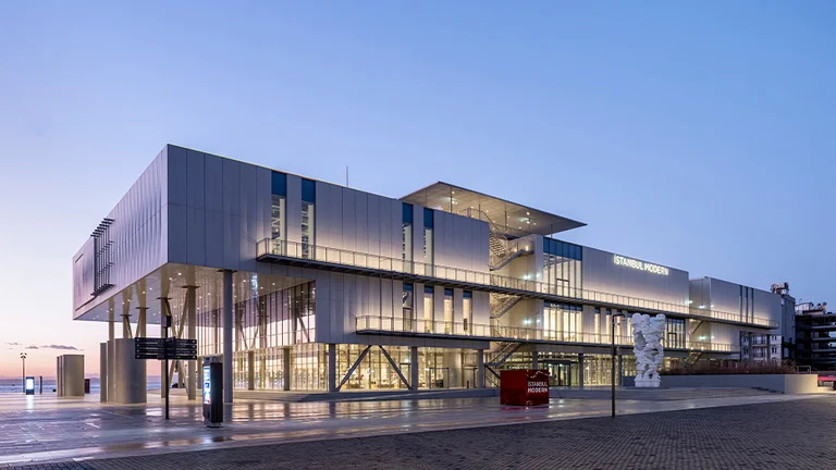 İstanbul Modern Museum at dusk with illuminated interiors, clear skies, and a sculpture out front.