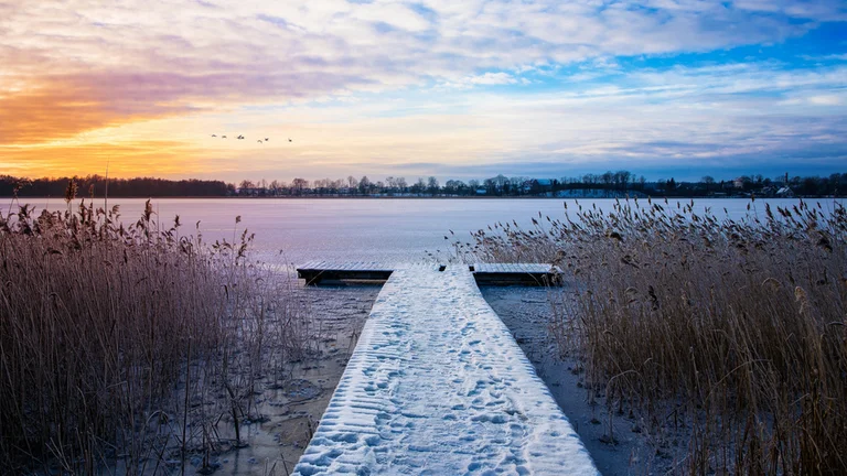 Snow-covered path leading to a frozen lake with reeds and a colorful sunrise.