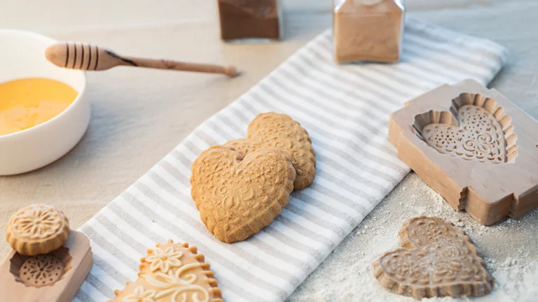 A cozy baking scene featuring heart-shaped cookies, honey, and decorative molds on a striped cloth with flour dust.