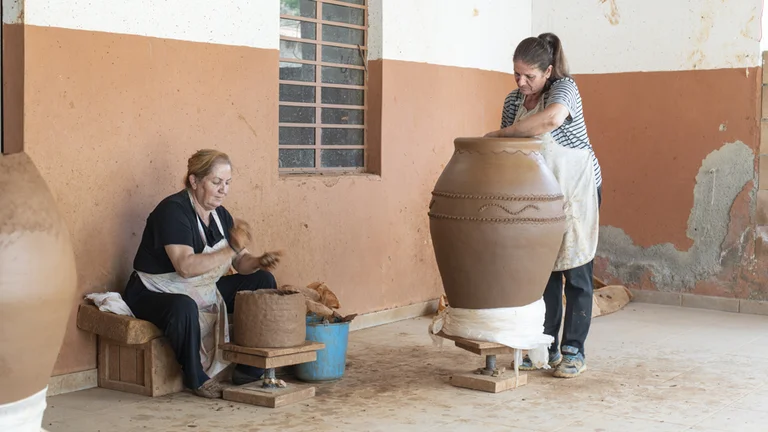 Two artisans shape clay pottery in a studio, one working on a large vase while the other prepares smaller pieces. Tools and materials are visible.