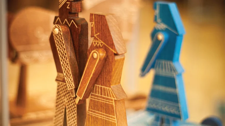 Two intricately carved wooden figures, a man and a woman, stand on a decorative base, alongside a blue figurine in the background.